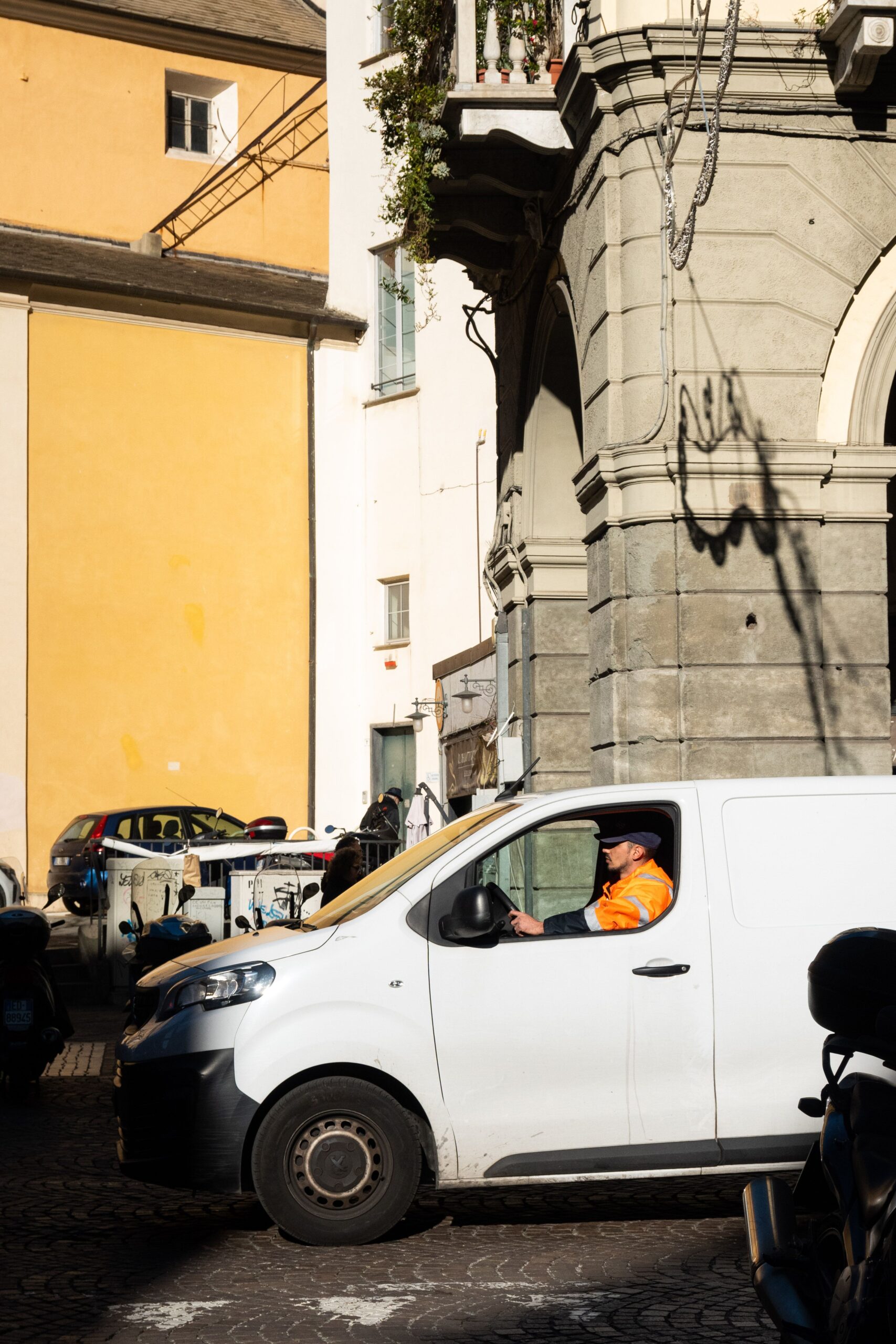 A white van driving through a sunlit street in Savona, Liguria, Italy.